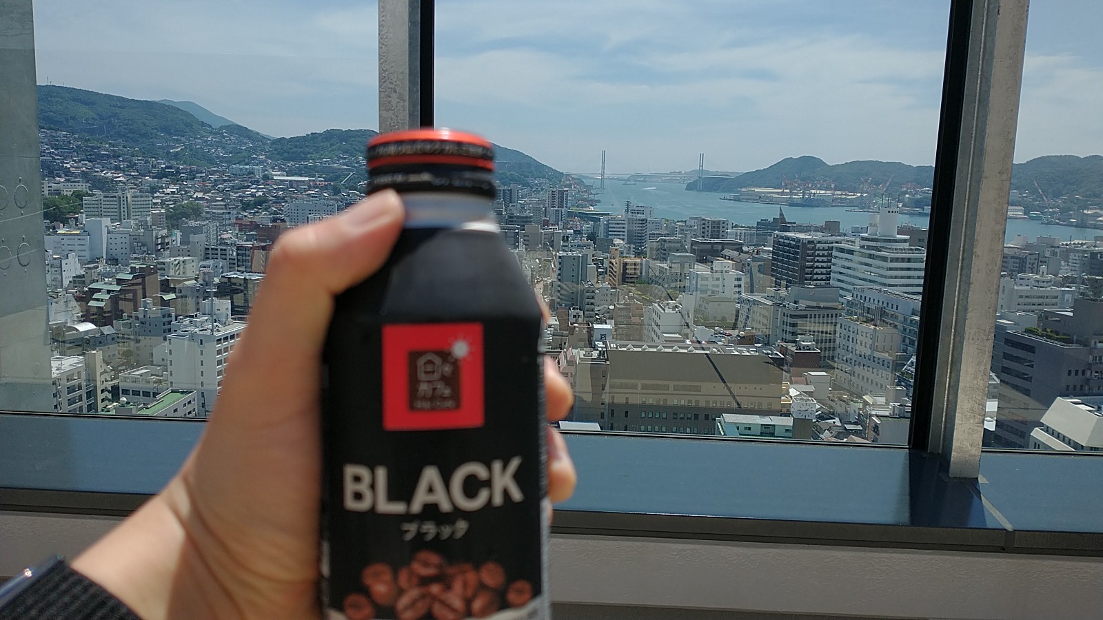 In the foreground, you can see a hand holding a bottle of coffee. That’s not what’s important, though. Beyond the window, you can see plenty of rooftops, and beyond that, a suspension bridge across the mouth of a harbour.