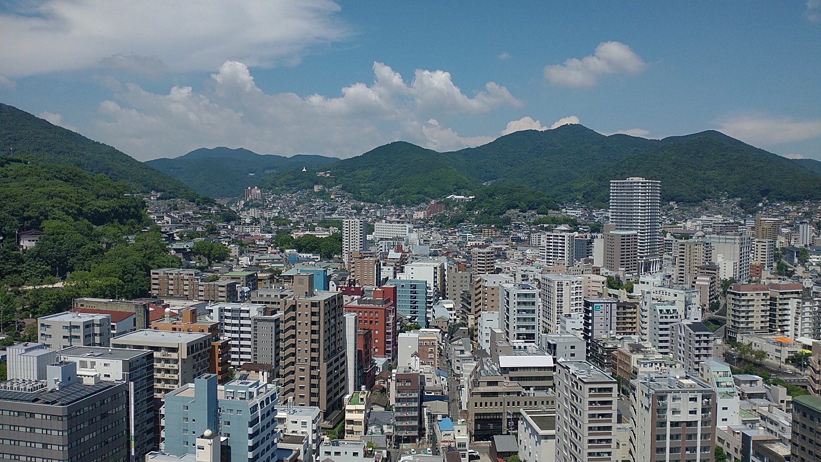 It’s a bright sunny day with white clouds and a light-blue sky. You can see plenty of buildings into the yonder, with mountains to the left and in the background of the picture. If you look carefully to the left, you can see Suwa Shrine, a noted “Power Spot” in Nagasaki City.