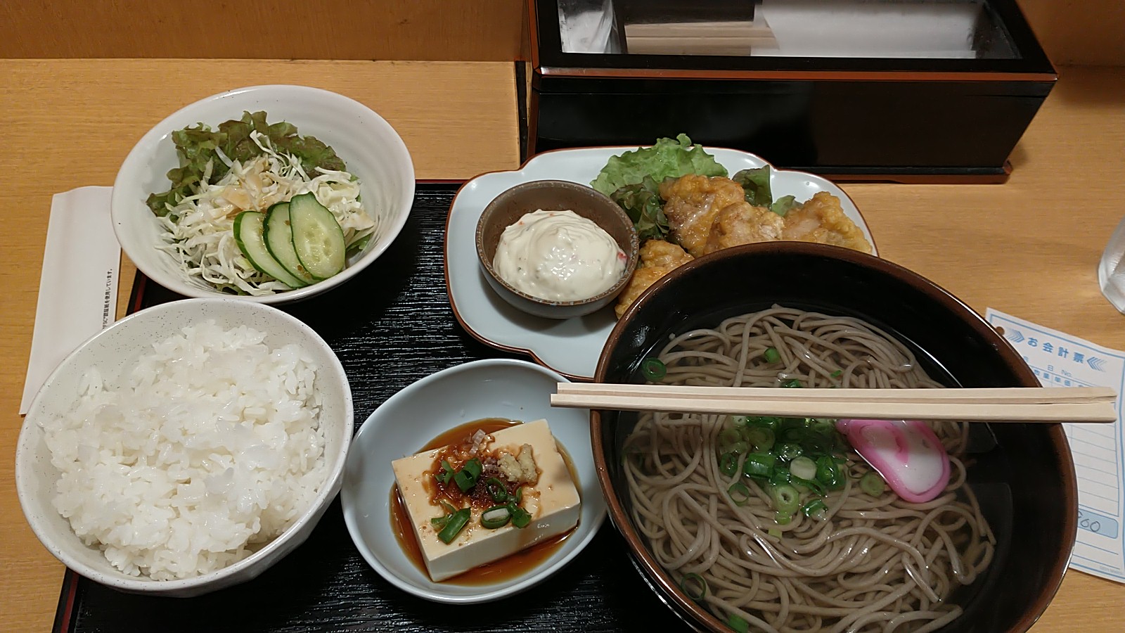 A tray of Japanese food. On the top left, we have a small salad, and on the right there is a dish with fried chicken and Japanese tartar sauce. At the bottom of the tray, there is a bowl of rice, a small dish with tofu, and a large bowl of soba noodles. 